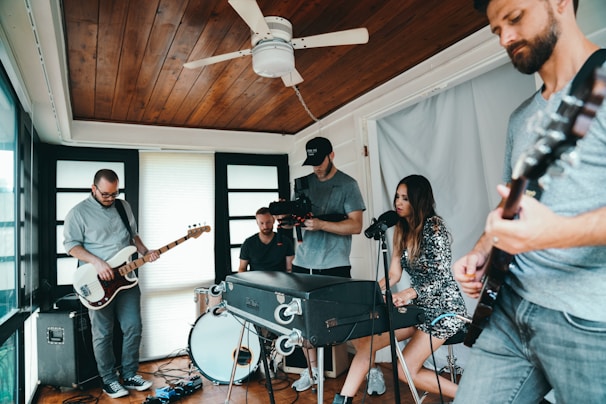 A group of musicians is performing indoors, with one person playing the bass guitar, another on an electric guitar, a drummer, and a singer seated at a piano. A videographer is capturing the scene. The room has wooden ceilings and large windows, creating a cozy and intimate setting.