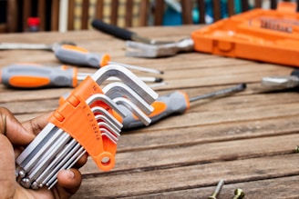 A set of hex keys with an orange holder is being held in a hand on a wooden table. Various other tools, including screwdrivers and a toolbox, are scattered on the table in the background.
