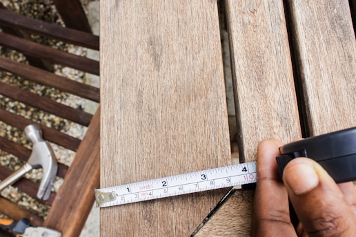 A close-up view of a person's hand holding a tape measure against a wooden surface. The tape measure extends to a marked length, and a hammer is seen in the background on a wooden slatted surface.