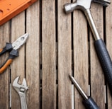 A collection of hand tools arranged on a wooden surface, including a hammer with a black handle, pliers with orange grips, an adjustable wrench, and a screwdriver.