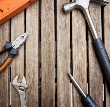 A collection of hand tools arranged on a wooden surface, including a hammer with a black handle, pliers with orange grips, an adjustable wrench, and a screwdriver.