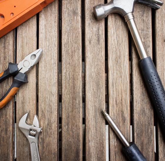 A collection of hand tools arranged on a wooden surface, including a hammer with a black handle, pliers with orange grips, an adjustable wrench, and a screwdriver.