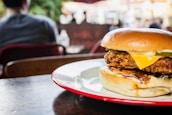 A large burger with a crispy patty, melted cheese, and layers of fresh vegetables sits on a white and red plate. The background softly blurs people and outdoor seating, suggesting a casual dining atmosphere.