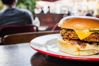 A large burger with a crispy patty, melted cheese, and layers of fresh vegetables sits on a white and red plate. The background softly blurs people and outdoor seating, suggesting a casual dining atmosphere.