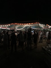 A group of people gathered under a tent adorned with string lights, creating an outdoor evening setting. The area is dimly lit, with people standing and mingling, suggesting a social event or festival.