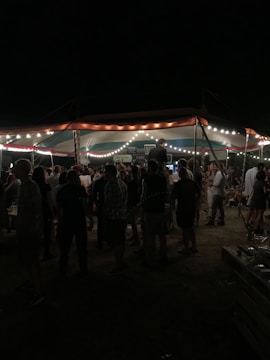 A group of people gathered under a tent adorned with string lights, creating an outdoor evening setting. The area is dimly lit, with people standing and mingling, suggesting a social event or festival.