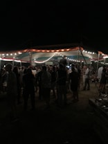 A group of people gathered under a tent adorned with string lights, creating an outdoor evening setting. The area is dimly lit, with people standing and mingling, suggesting a social event or festival.