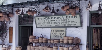 A quaint shop facade displays baskets and a sign in Greek. The exterior is adorned with hanging lights, garlic bulbs, and woven items. The atmosphere evokes a traditional, rustic charm.