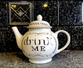 A white ceramic teapot with a decorative lid and the inscription 'drink ME' on its side. It is placed on a speckled stone countertop against a dark, patterned tile backsplash.
