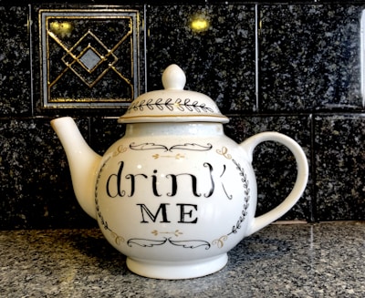 A white ceramic teapot with a decorative lid and the inscription 'drink ME' on its side. It is placed on a speckled stone countertop against a dark, patterned tile backsplash.