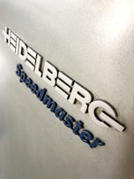 Metallic surface with embossed lettering that spells 'Heidelberg Speedmaster'. The letters are in a blue and white color scheme on a textured silver background.