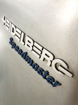 Metallic surface with embossed lettering that spells 'Heidelberg Speedmaster'. The letters are in a blue and white color scheme on a textured silver background.