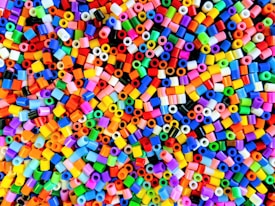 A multitude of small, colorful plastic beads scattered in a chaotic pattern. The beads are cylindrical in shape and appear in a wide array of vibrant colors including red, blue, green, yellow, purple, and more.