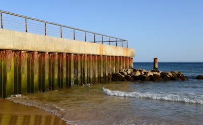 A concrete seawall extends into the ocean, with rust and green algae growth on its surface. Waves gently lap against the shore, and the clear blue sky meets the horizon.