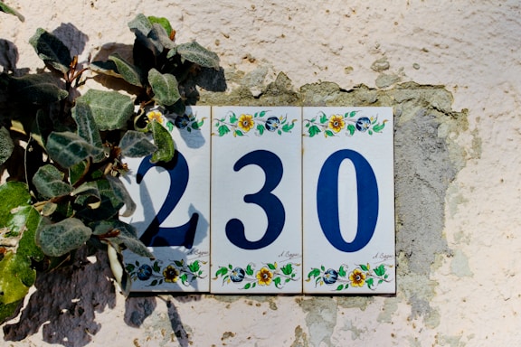 A decorative house number plaque displaying the numbers 230 in blue, set against a textured wall with green leaves partly covering the first digit. Each tile of the plaque features colorful floral designs at the top and bottom.