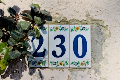 A decorative house number plaque displaying the numbers 230 in blue, set against a textured wall with green leaves partly covering the first digit. Each tile of the plaque features colorful floral designs at the top and bottom.