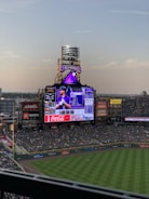 A baseball stadium with a large electronic scoreboard displaying player information. The seating area is moderately filled with spectators. The field is well-maintained, and evening sky is visible.
