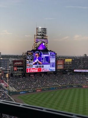 An outdoor sports stadium with a massive digital LED scoreboard lit up during evening game.