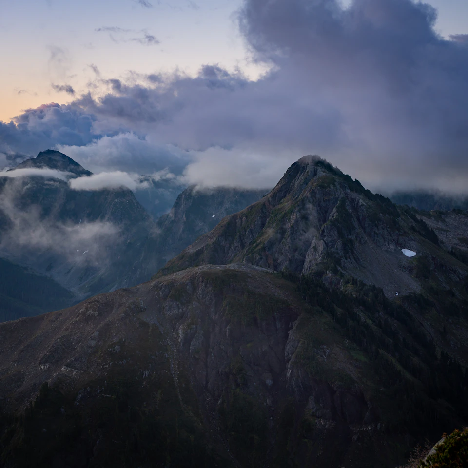 The image depicts a majestic mountain range with a cloudy sky above, creating an atmospheric and dramatic backdrop for the scene. The mountains are covered in lush green vegetation, adding depth to the landscape. The perspective is from a high vantage point, providing a bird's eye view of the mountains and their surroundings.