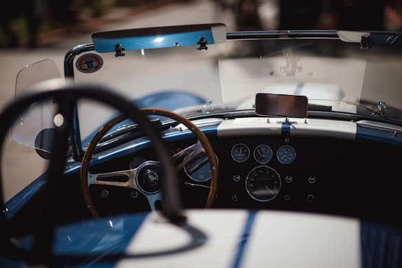 An interior view of a classic car, focusing on the dashboard with a wooden-rimmed steering wheel. The dashboard features multiple analog gauges and a mounted electronic device resembling a smartphone. The windshield displays a small sticker, and the color scheme is predominantly blue with white racing stripes.