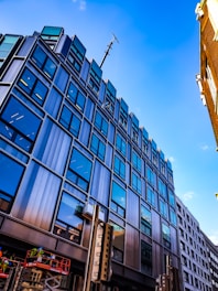 A modern multi-story building with reflective glass windows set against a clear blue sky. The structure features a sleek design with steel and glass, emphasizing an urban architectural style. Construction workers are visible on a raised platform at the bottom left, wearing safety gear.