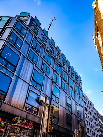 A modern multi-story building with reflective glass windows set against a clear blue sky. The structure features a sleek design with steel and glass, emphasizing an urban architectural style. Construction workers are visible on a raised platform at the bottom left, wearing safety gear.