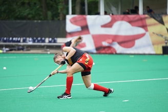 A field hockey player in red shorts and a black top is actively swinging a hockey stick towards a ball on a green turf. The athlete is wearing red knee-length socks and black shoes. In the background, there are benches and a large colorful banner with abstract shapes. Another player is faintly visible, bending down in the distance.
