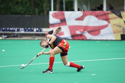 A field hockey player in red shorts and a black top is actively swinging a hockey stick towards a ball on a green turf. The athlete is wearing red knee-length socks and black shoes. In the background, there are benches and a large colorful banner with abstract shapes. Another player is faintly visible, bending down in the distance.