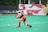 A field hockey player in red shorts and a black top is actively swinging a hockey stick towards a ball on a green turf. The athlete is wearing red knee-length socks and black shoes. In the background, there are benches and a large colorful banner with abstract shapes. Another player is faintly visible, bending down in the distance.