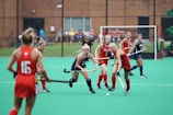 A group of female field hockey players in red and blue uniforms are actively playing a match. The action is focused on a player in red closely guarding another player in blue who is attempting to control the ball. Spectators are visible in the background, seated along the fenced perimeter of the field.