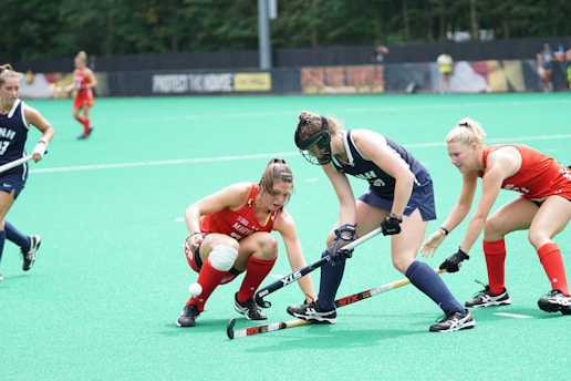 A group of field hockey players is intensely competing for the ball on a grassy field. Two players in red uniforms are challenging two players in dark blue uniforms. The athletes are using sticks to control the ball, with one central player actively engaged in controlling it. The background features a vibrant green playing surface and trees.