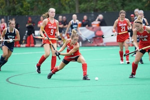 A group of female field hockey players are actively participating in a game on a green turf field. They are wearing red and blue sports uniforms, with some players from the Maryland team. One player is prominently attempting to hit the ball with her hockey stick, focused and poised. Spectators can be seen in the background.