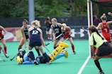 A field hockey match with players actively competing near the goal. Two teams in blue and red jerseys are involved, with a player in blue attempting a move as the goalkeeper lies on the ground in protective gear. A referee in a yellow and black jersey is observing the play closely. Lush greenery and a banner can be seen in the background.