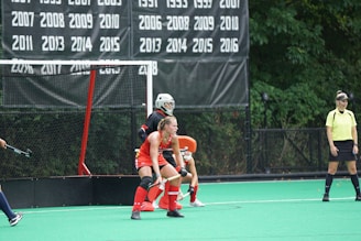 A field hockey game in progress with a player wearing a red jersey and protective gear positioned defensively in front of the goal. Another player, likely a goalkeeper, is outfitted in all black and is also in front of the goal. An official wearing a yellow shirt and black skirt stands nearby, observing the play. The backdrop features a list of years on a black banner, signifying past achievements or championships.
