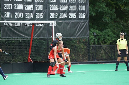 A field hockey game in progress with a player wearing a red jersey and protective gear positioned defensively in front of the goal. Another player, likely a goalkeeper, is outfitted in all black and is also in front of the goal. An official wearing a yellow shirt and black skirt stands nearby, observing the play. The backdrop features a list of years on a black banner, signifying past achievements or championships.