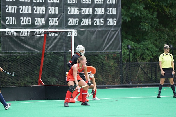 A field hockey game in progress with a player wearing a red jersey and protective gear positioned defensively in front of the goal. Another player, likely a goalkeeper, is outfitted in all black and is also in front of the goal. An official wearing a yellow shirt and black skirt stands nearby, observing the play. The backdrop features a list of years on a black banner, signifying past achievements or championships.