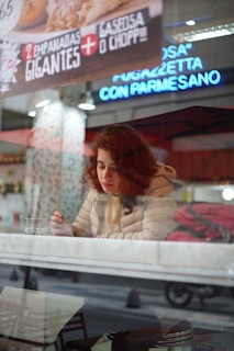 A person with curly red hair is sitting inside a cafe or restaurant, looking down while holding a utensil. They are wearing a beige quilted jacket. There is a glass cup on the table in front of them. The scene is viewed through a window with reflections of neon signs and a street outside. Above, a sign advertises '2 empanadas gigantes + gaseosa o chopp'.