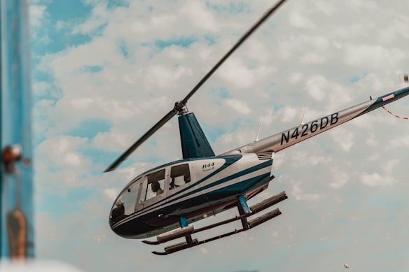 A helicopter is captured mid-flight against a backdrop of a cloudy sky. The helicopter has a blue and white design, with the registration number N426DB visible on its tail. The scene suggests an outdoor, possibly open-air location.