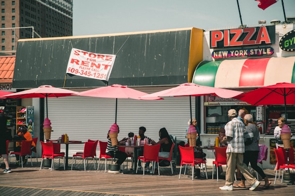 A boardwalk scene with people sitting under bright pink umbrellas at red tables. Behind them is a storefront with a sign indicating it's for rent. To the right, there is a pizza place with colorful signage including 'New York Style' text. People are walking past, and large ice cream-shaped trash bins are visible.