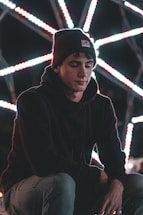 A person wearing a beanie and hoodie appears contemplative against a backdrop of bright, intersecting lines of light that create a geometric pattern.