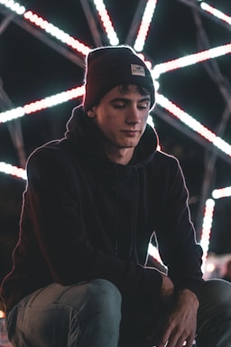 A person wearing a beanie and hoodie appears contemplative against a backdrop of bright, intersecting lines of light that create a geometric pattern.