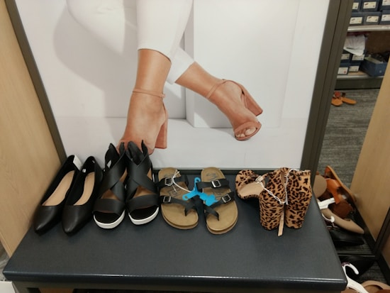 Various types of women's shoes are displayed on a bench, including black flats, black platform sandals, beige sandals with buckles, and leopard print high heels. Behind the shoes, a photo features a close-up of legs wearing beige high-heeled sandals.