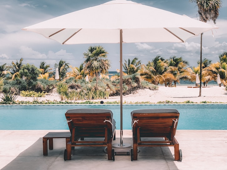 Two wooden lounge chairs with cushions are positioned by a swimming pool facing a sandy beach. A large white umbrella provides shade over them. In the background, there are tall palm trees and lush greenery, set against a partly cloudy sky.