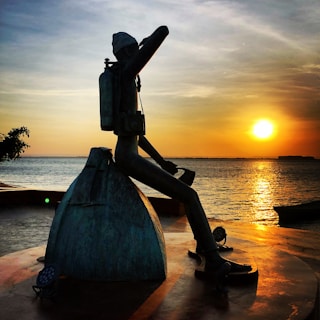 A silhouette of a person sitting on a backpack overlooks a calm body of water during a vibrant sunset. The figure is wearing a hat and carrying a camera around their neck, suggesting they are a traveler or photographer. The sun casts a warm golden glow across the scene, reflecting off the water.