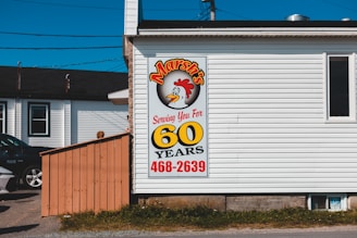 A building with white siding featuring a large vertical sign. The sign has a cartoon chicken and reads 'Marsh's' with the message 'Serving You For 60 Years'. A phone number, 468-2639, is displayed in red at the bottom. A parked car and another building are visible in the background.