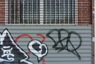A metal roller door features various graffiti including a stylized heart and abstract shapes in black, white, and red. A brick wall and barred windows are visible above the door.