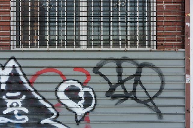 A metal roller door features various graffiti including a stylized heart and abstract shapes in black, white, and red. A brick wall and barred windows are visible above the door.