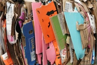 A stack of various flattened cardboard boxes and packaging is piled together. The boxes are of different sizes and colors, including bright orange, pink, blue, and green. Some have printed text and images, such as black bats on an orange background, indicating a Halloween theme.