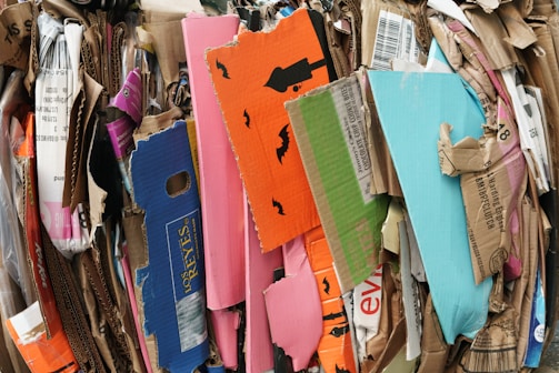 A stack of various flattened cardboard boxes and packaging is piled together. The boxes are of different sizes and colors, including bright orange, pink, blue, and green. Some have printed text and images, such as black bats on an orange background, indicating a Halloween theme.