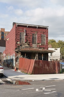 A derelict, red brick building showing signs of decay, with boarded-up windows and faded, peeling paint. The structure is covered in various graffiti, and its worn-out, rusted roof is supported by a rickety framework. A red metal fence encircles the property, adding a sense of abandonment. The pavement around the building is chipped, and there is a puddle of water nearby, with a road in the foreground.